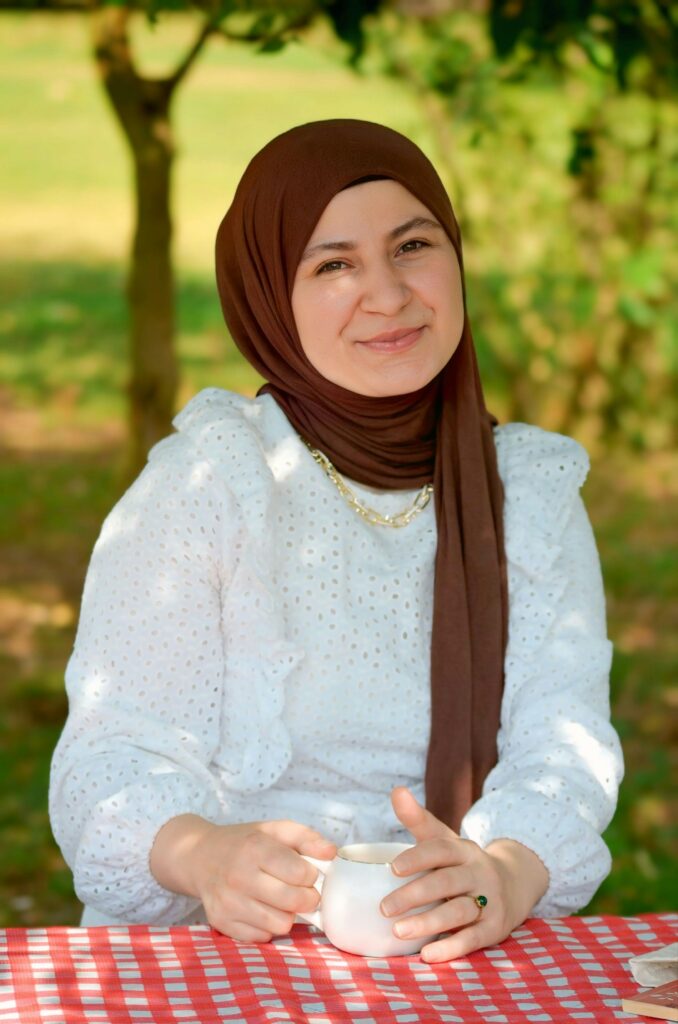 Smiling woman in hijab enjoying coffee outdoors on a sunny day, at a picnic table.